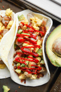 A white tortilla filled with eggs, sausage, bacon, and avocado, and topped with Sriracha.