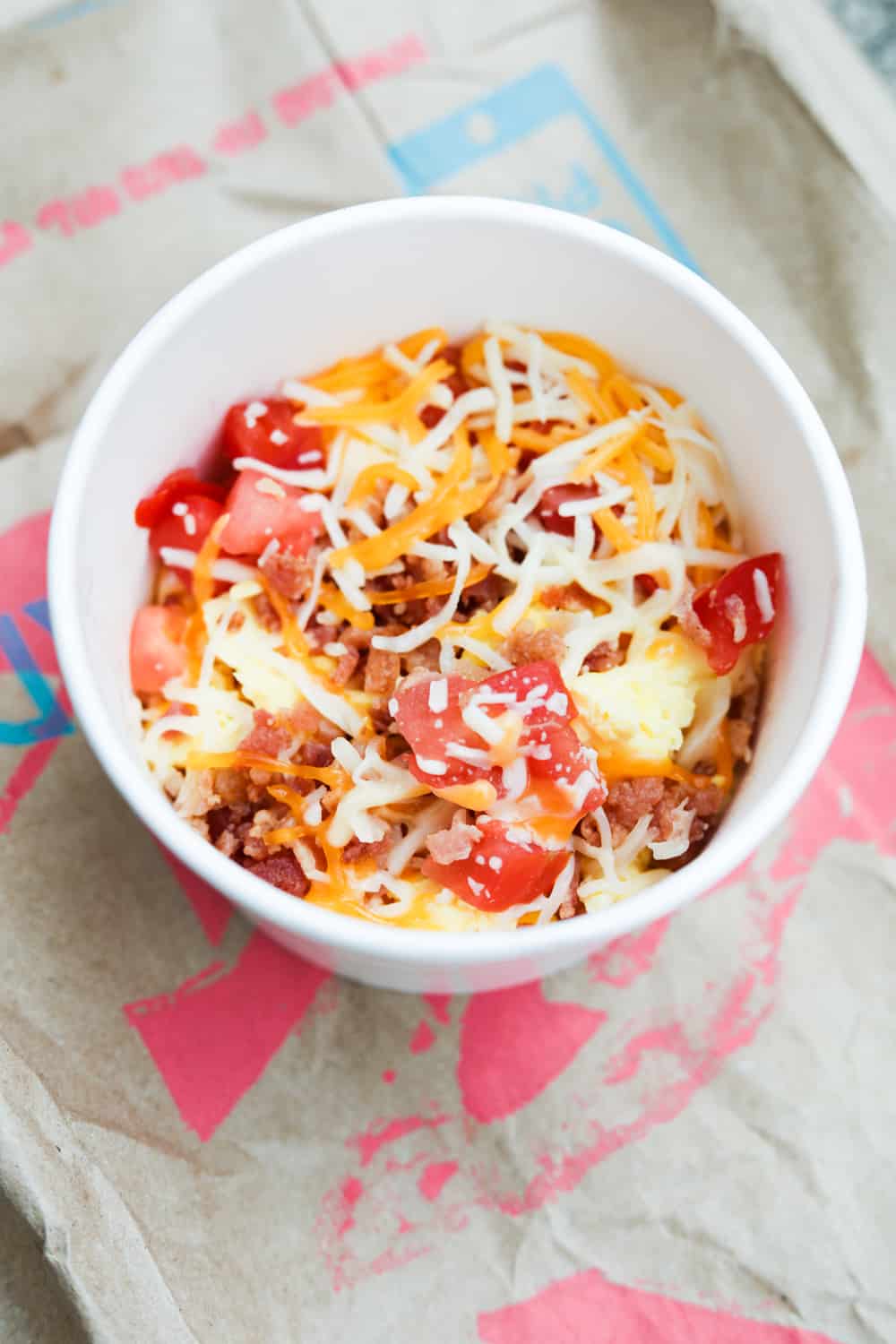 A white cup with shredded cheese, egg, tomatoes, and bacon in it. The cup is on a brown paper bag.