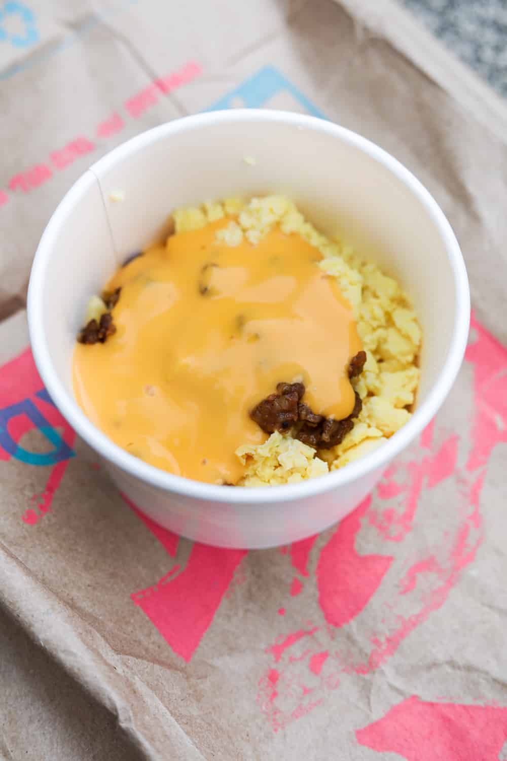 A white cup filled with egg, sausage crumbles, and nacho cheese sauce. The cup is on a brown paper bag.