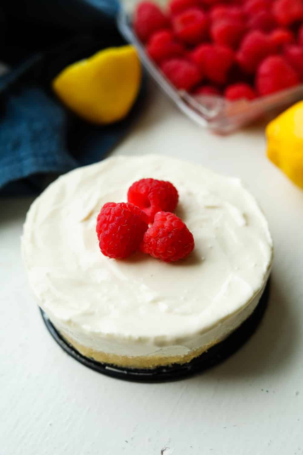 Cheesecake topped with raspberries. There's a blue towel, lemons, and raspberries behind the cheesecake.