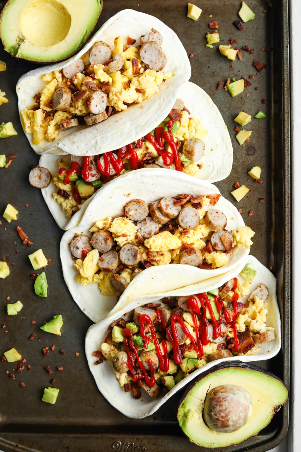 Breakfast tacos on a baking sheet. The tacos are filled with eggs, bacon, sausage. Some of the tacos have avocado and Sriracha on them as well.