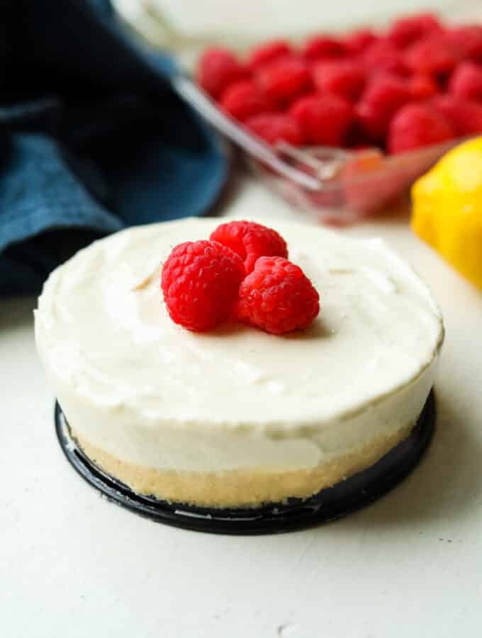 Cheesecake topped with cheesecake with raspberries and lemons set behind it.