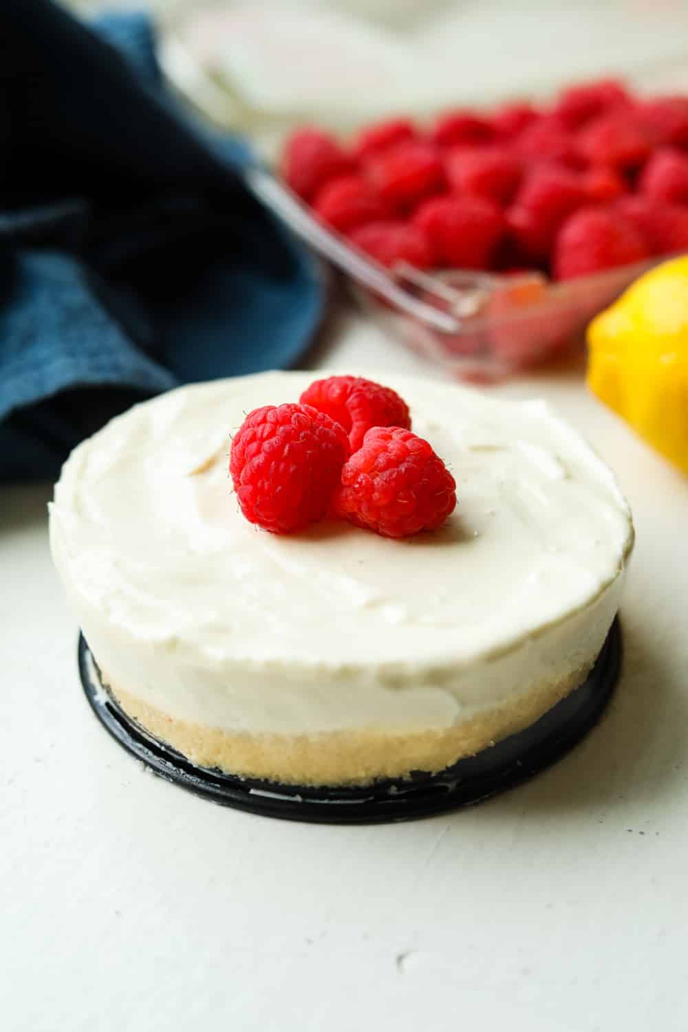 Cheesecake topped with cheesecake with raspberries and lemons set behind it.