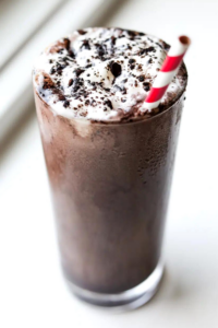 A glass filled with an oreo protein shake topped with whipped cream. There is a white and red ribbon straw in the glass and the glass is on a white counter.