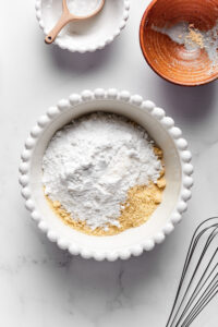 A medium sized white bowl with almond flour and confectioners erythritol in it. The balls on a white counter and there is a whisk in front of it and an empty small brown bowl behind it and an empty small white ball next to that.