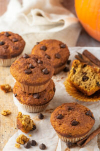 Two pumpkin muffins stacked on top of one another with two more muffins and one muffin with cut in half on a piece of parchment paper on a white counter.