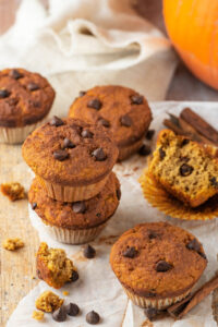 Two pumpkin muffins stacked on top of one another with two more muffins and one muffin with cut in half on a piece of parchment paper on a white counter.