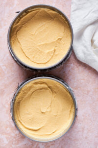 To springform pans on a pink counter filled with vanilla cake batter.