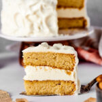 A slice of vanilla cake with vanilla frosting and a serving spatula and a white counter. Behind it is the entire cake with a slice taken out of it on a cake platter.