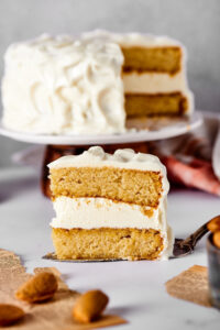 A slice of vanilla cake with vanilla frosting and a serving spatula and a white counter. Behind it is the entire cake with a slice taken out of it on a cake platter.