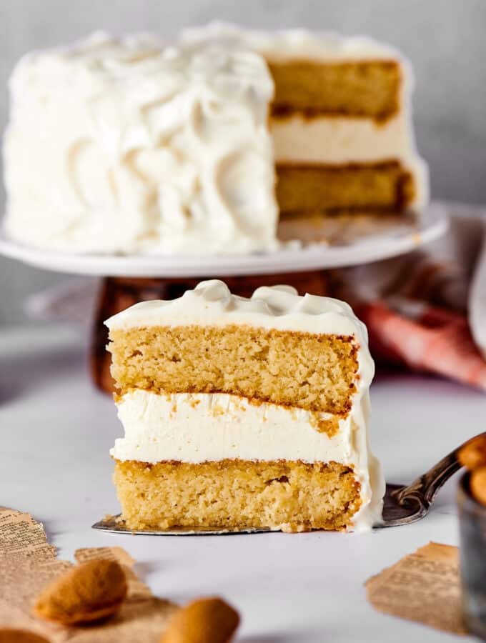 A slice of vanilla cake with vanilla frosting and a serving spatula and a white counter. Behind it is the entire cake with a slice taken out of it on a cake platter.