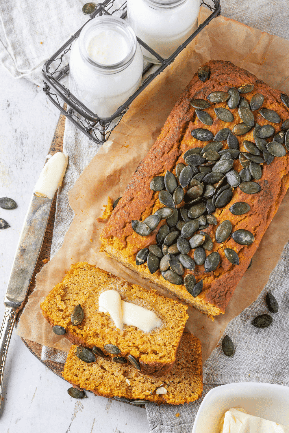 A loaf of keto pumpkin bread on top of a piece of brown parchment paper on a gray tablecloth. There are two slices of pumpkin bread cut out line face up in front of the loaf. Next to them is a knife with butter on it and in front of that is a metal crate with two glass jars of milk.