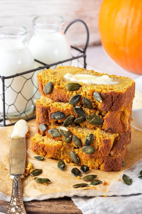 Four slices of pumpkin bread on top of one another in a stack on a piece of brown parchment paper on a gray tablecloth. There is a knife with butter on it to the left of the bread and behind it is a wire holder with two glass jars of milk in it. To the back right is part of a pumpkin.