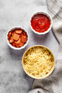 A bowl of shredded mozzarella cheese, a small white bowl of pepperoni, and a small white bowl of pizza sauce all on a grey counter.