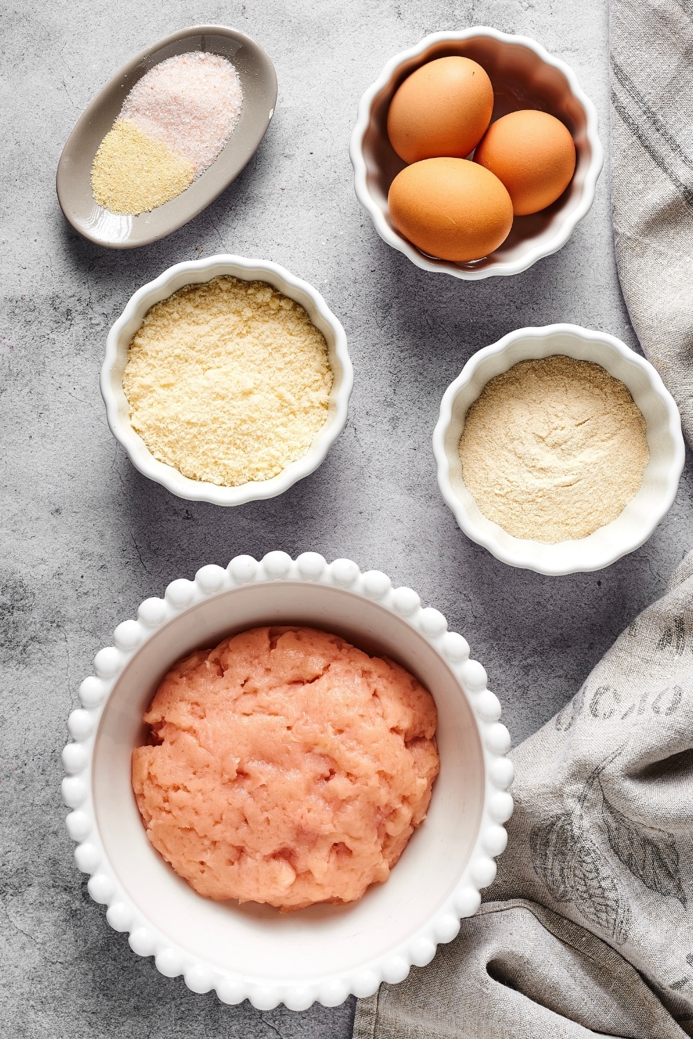 A white dish with ground chicken in it, a small white bowl with protein powder, a small white bowl with parmesan cheese, a white bowl with three eggss, an oval dish with garlic salt and onion powder all on a grey counter.