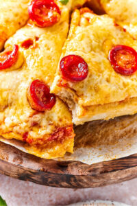 A slice of chicken crust pizza with cheese and pepperoni laying on the side of another slice on a piece of parchment paper on a wooden board.