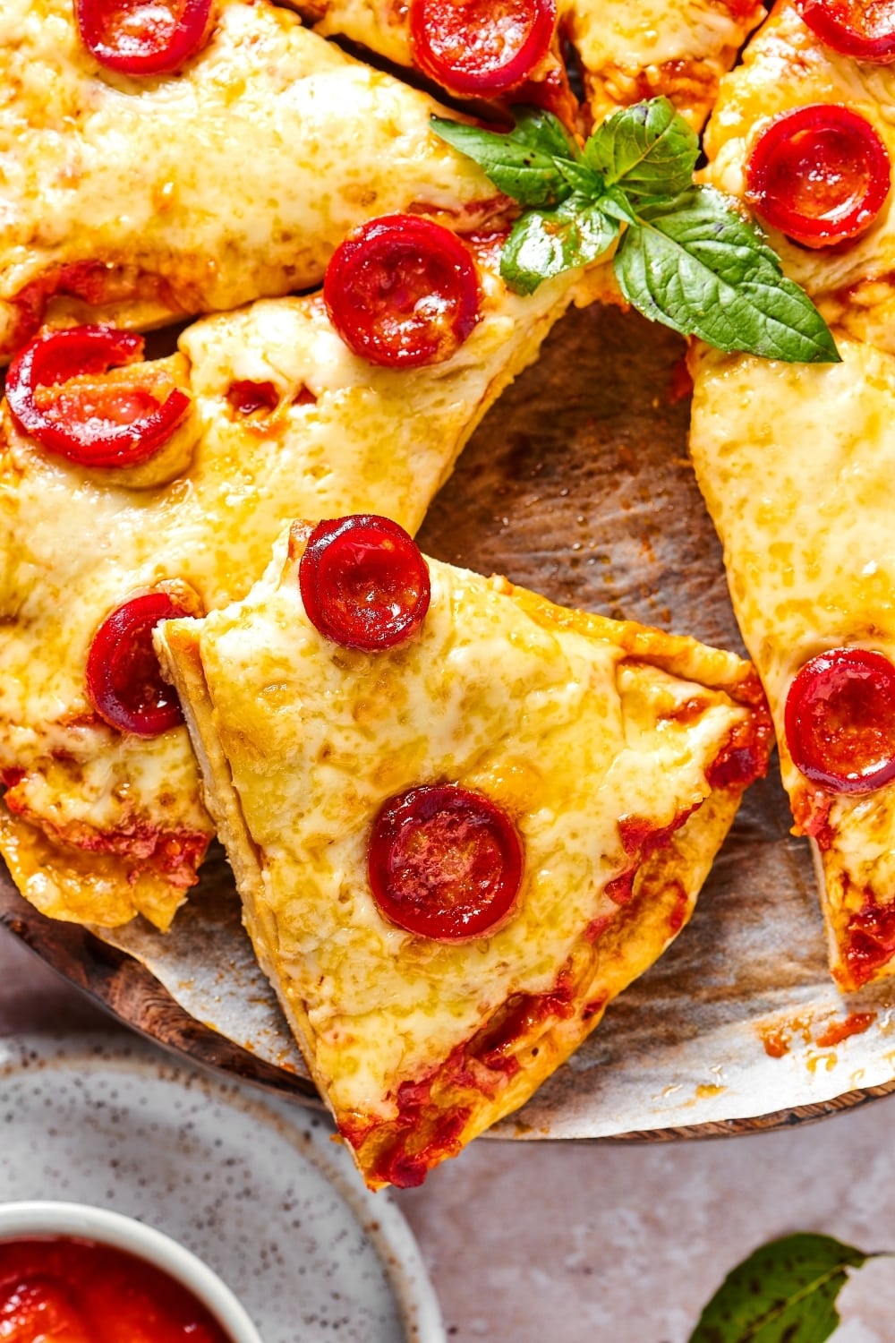 A slice of cheese and pepperoni pizza with a bite out of the front on top of another piece on a piece of parchment paper. Part of the rest of the pizza is on the board.