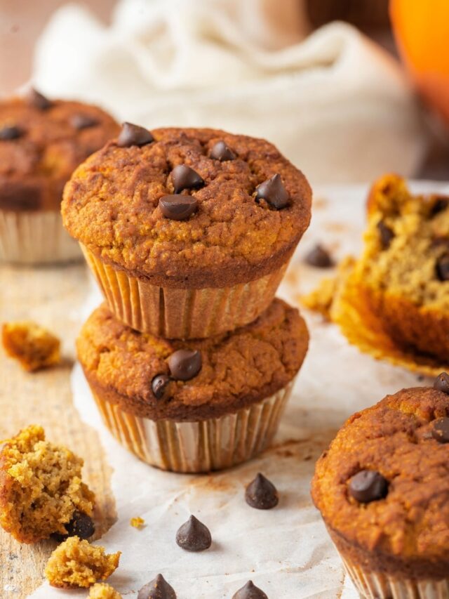 Two pumpkin muffins stacked on top of one another on a piece of parchment paper on a wooden counter. There are a couple of chocolate chips in front of it and a piece of pumpkin muffin on the wooden counter next to the parchment paper.