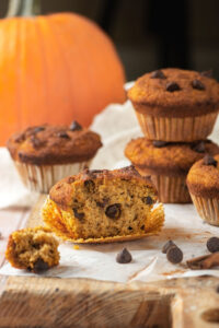 A pumpkin muffin with the front missing from it on a muffin wrapper on a piece of parchment paper on a wooden counter. Behind it is another pumpkin muffin with two pumpkin muffins on top of each other to the right of that. Behind those muffins is a pumpkin.