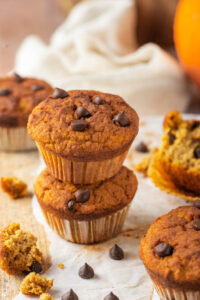 Two pumpkin muffins stacked on top of one another on a piece of parchment paper on a wooden counter. There are a couple of chocolate chips in front of it and a piece of pumpkin muffin on the wooden counter next to the parchment paper.