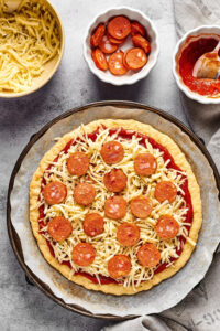 An uncooked chicken crust pizza with cheese and pepperoni on it on a piece of parchment paper on a pizza tray. Behind it is a white bowl of pepperoni, a white bowl of pizza sauce, and a white bowl of shredded mozzarella cheese.