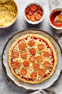 An uncooked chicken crust pizza with cheese and pepperoni on it on a piece of parchment paper on a pizza tray. Behind it is a white bowl of pepperoni, a white bowl of pizza sauce, and a white bowl of shredded mozzarella cheese.