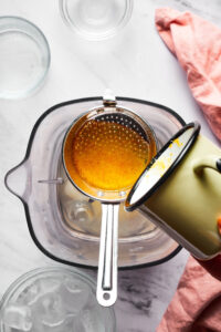 A cup pouring orange syrup through a mash Shiva into a blender with margarita ingredients in it. The blender is on a white counter and behind it as part of a cup of ice and in front of it is two empty glass bowls.