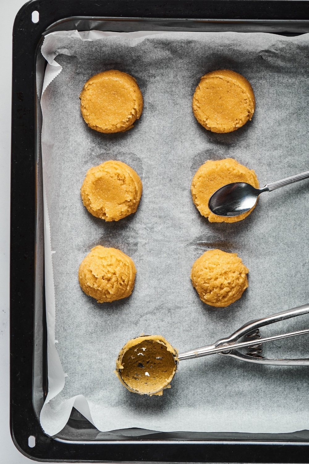 A baking tray line with parchment paper with two rolls of three shortbread cookies on it. In the right roll a spoon is on top of the middle cookie and behind the rose in the middle of them is a cookie scooper.