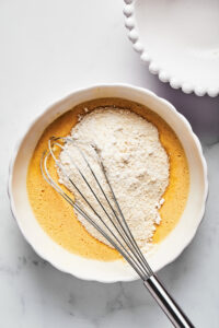 A large white bowl with wet poundcake ingredients and pound cake ingredients on top of that. There is a whisk in the bowl and the bowl is on a white counter.