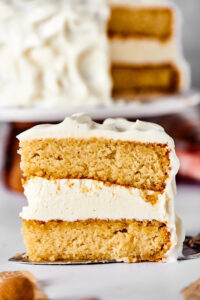 A slice of vanilla cake with vanilla frosting being held by a cake spatula. Behind it is the vanilla cake with the slice missing.