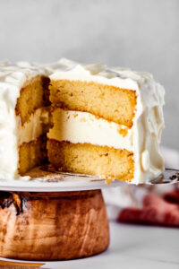 A slice of cake being pulled from a vanilla cake with vanilla frosting that is on the cake stand.