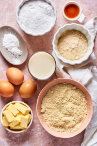 Ingredients for keto cake. There is a medium bowl with almond flour, a bowl with sticks of butter, three eggs in front of that, a cup of milk next to the eggs, a small bowl of coconut flour, a small bowl of vanilla extract, an oval dish of baking powder, and a small white dish of confectioner erythritol.