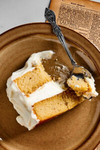 A slice of vanilla cake on top of a brown plate on a piece of newspaper. A spoon is rusting on the edge of the plate with the head of the spoon right next to the cake with a piece of it on it.