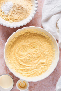 A white bowl with vanilla cake batter in it. Behind that is part of another white ball with the dry ingredients for the vanilla cake batter in it. Both bowls are on a pink counter.