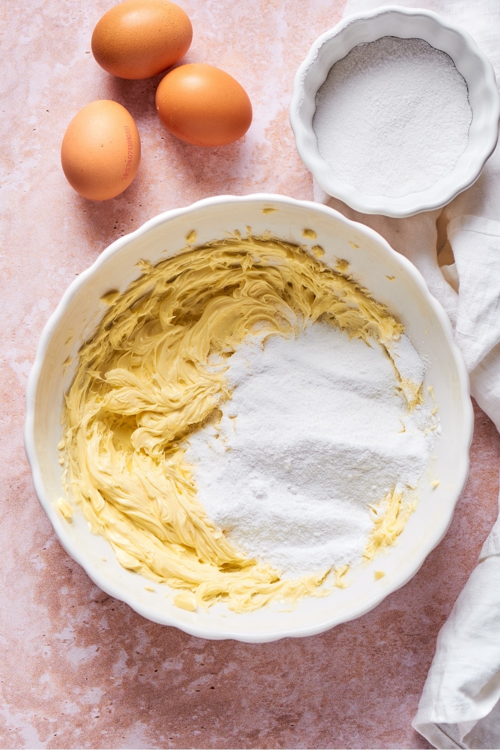 A white bowl filled with whipped cake batter with some powdered erythritol on top of the right side of it. Behind the bowl is a small bowl of powdered erythritol and three eggs to the left of it.