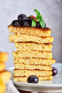 Pieces of eight cream cheese pancakes stacked on top of one another on a white plate. There are blueberries on the top piece and two blueberries on the white plate.