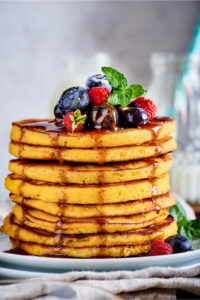 I ate cream cheese pancakes on top of one another on a white plate. There is syrup dripping down the pancakes and there are blueberries and raspberries on the top pancake.