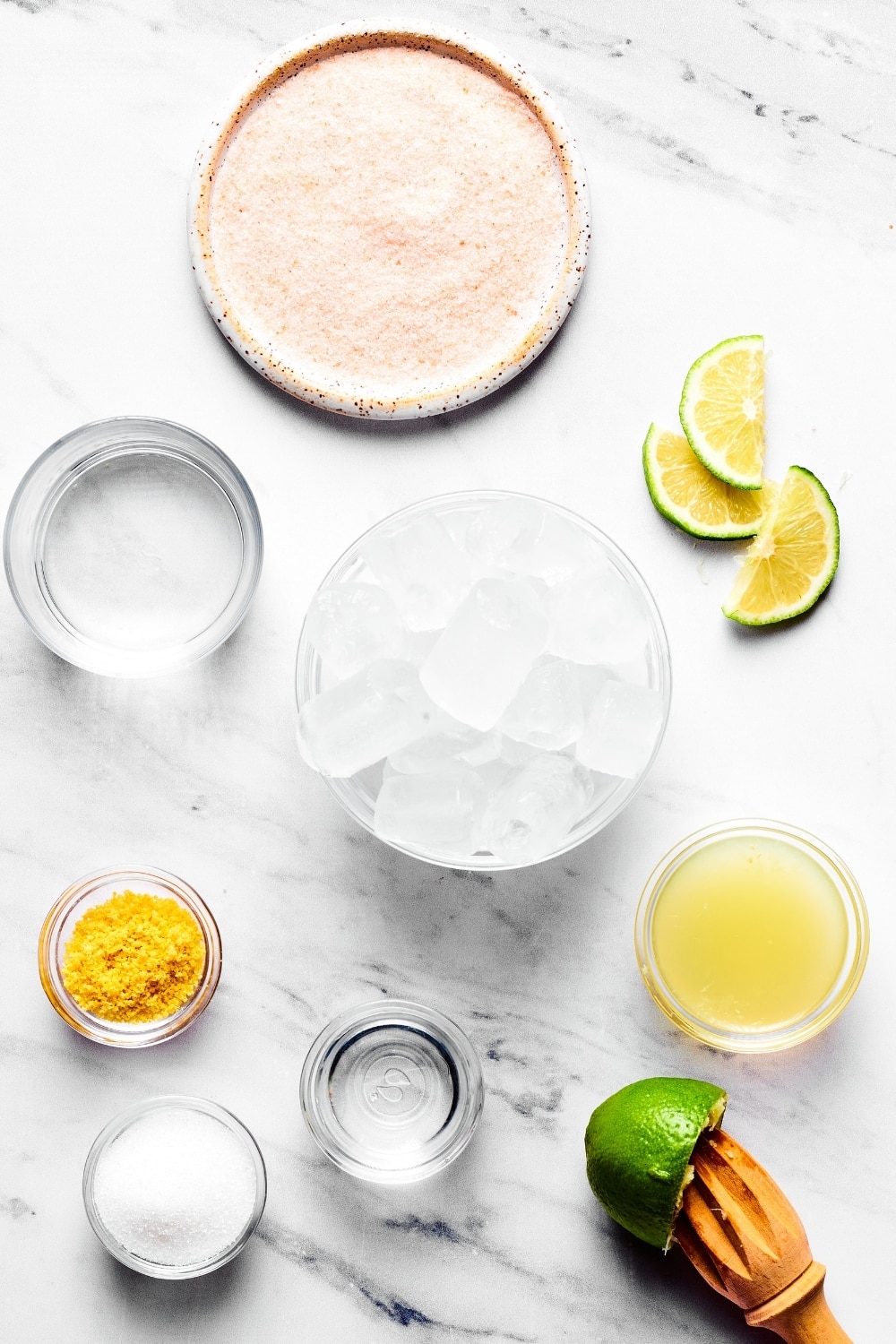 Keto margarita ingredients on a white counter. There is a small bowl of erythritol, small bowl of orange zest, a small cup of tequila, a bowl of ice, a small cup of water, if you wind wedges, and a plate of salt.