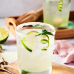 Glass of margarita with a lime wedge in it on a wooden board. There are a few lime wedges in front of it and behind the glass is a wooden serving tray with another glass of margarita on it.