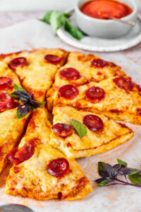 A chicken pizza crust cut into slices with cheese and pepperoni on it on a piece of parchment paper.