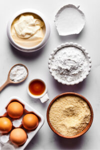 A medium bowl with almond flour, a carton of four eggs to the left of that, a small bowl of vanilla extract and small spoon of baking powder in front of the eggs, a bowl of confectioners erythritol in front of the almond flour, in front of that a white leaf bowl with melted coconut oil, and next to that a medium white bowl with cream cheese. Everything is on a white counter.