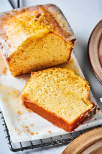 A loaf of poundcake on a piece of white parchment paper on a black wire rack that is on a white counter. A slice of pound cake from the front of the loaf is lying flat in front of the entire pound cake.