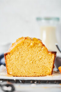 A vanilla poundcake loaf with a slice out of the front of it on a piece of parchment paper on a black wire rack set on a white counter.