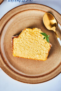 Hey slice of pound cake line flat on a brown plate that is on a white counter. There is a gold spoon on the plate at the top of the poundcake.