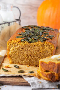 A pumpkin bread loaf on a piece of parchment paper on a wood board. There is part of two slices from the bread in front of it and there is part of a pumpkin behind it.