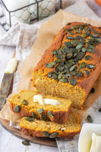 A loaf of pumpkin bread and a piece of parchment paper on a and a tablecloth that is on a wood board on the white counter. There are two slices of pumpkin bread line against one another in front of the loaf.