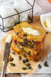 A gray tablecloth with a piece of brown parchment paper on it with four slices of pumpkin bread stacked on top of one another on that. There's a slab of butter on the top piece of pumpkin bread and a knife is next to the stack with butter on it. Behind it is part of a white dish with butter next to that a wire holder with two glass jars of milk in it.