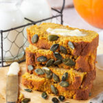 Four slices of pumpkin bread stack on top of one another on a piece of parchment paper on a gray table cloth. There are some pumpkin seeds in front of the slices and there is a knife to the left of them with butter on it. Behind the stack of pumpkin bread is of wire holder with two glass jars of milk.