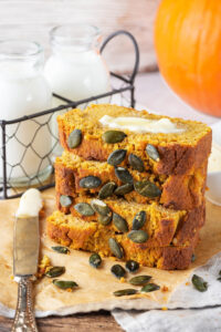 Four slices of pumpkin bread stack on top of one another on a piece of parchment paper on a gray table cloth. There are some pumpkin seeds in front of the slices and there is a knife to the left of them with butter on it. Behind the stack of pumpkin bread is of wire holder with two glass jars of milk.