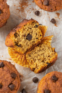 A pumpkin muffin on the muffin wrapper on a piece of parchment paper. The muffin is broken in half with the inside facing up and there are parts of other pumpkin muffins surrounding it.